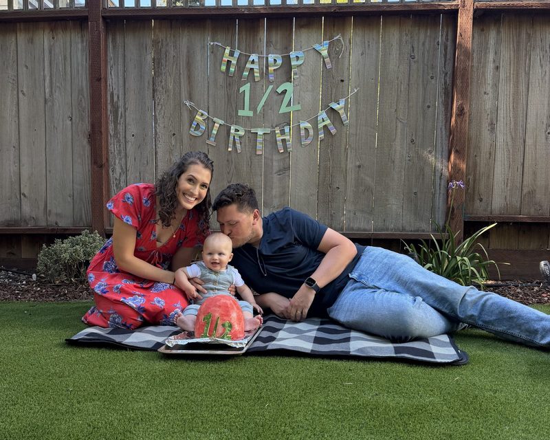 Taylor, his wife, and baby Clark on a picnic blanket in the backyard under a Happy Half Birthday banner