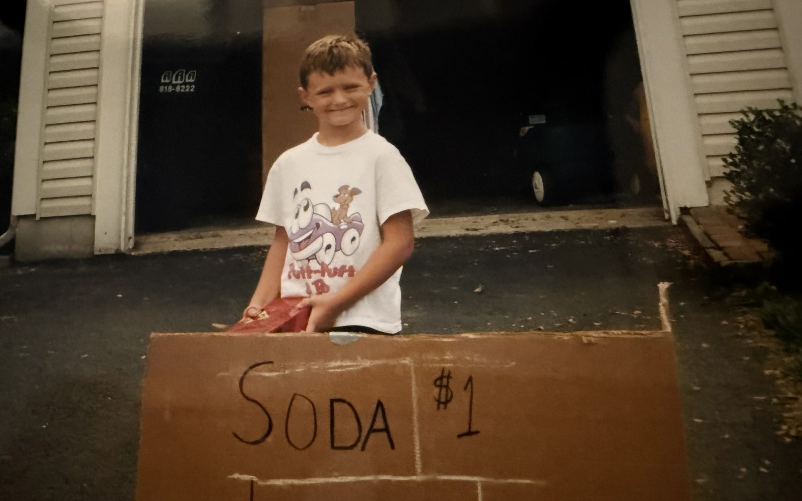 Taylor at age seven behind a hand-drawn cardboard sign in a driveway reading Soda one dollar, Lemonade twenty-five cents